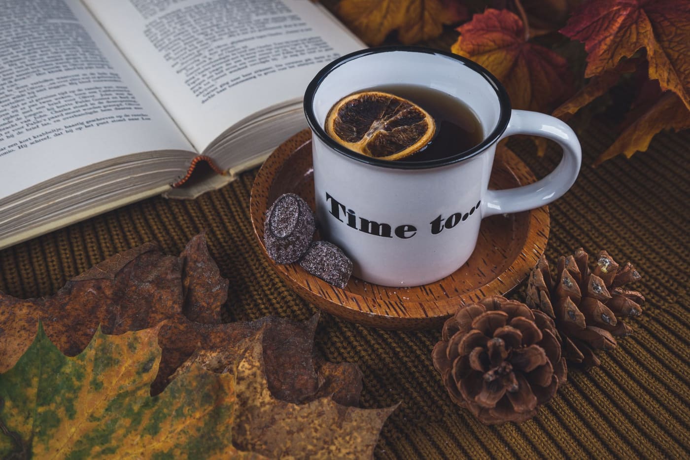 A mug of tea with a dried lemon slice beside an open book and autumn leaves