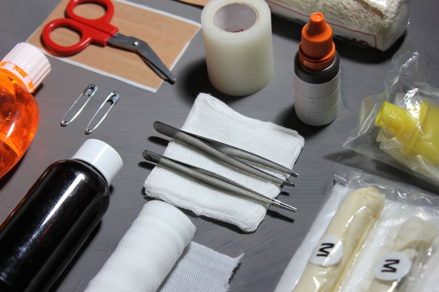 Gauze, tweezers, tape, scissors, gloves and bottles laid out on a dark table