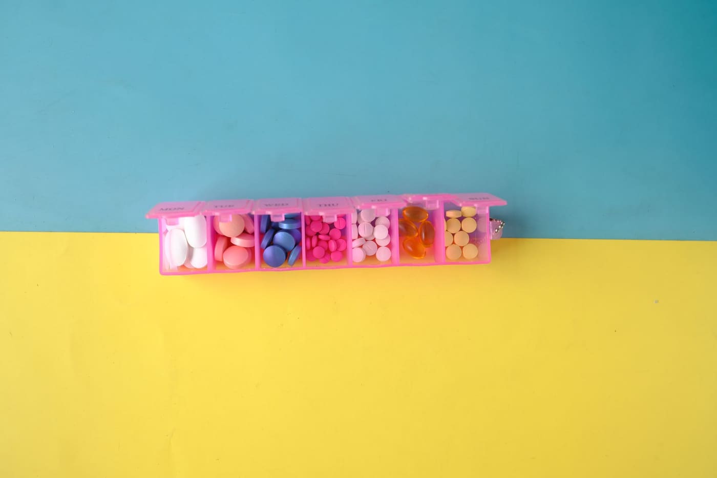 A pink weekly pill organizer with assorted tablets on a blue and yellow background